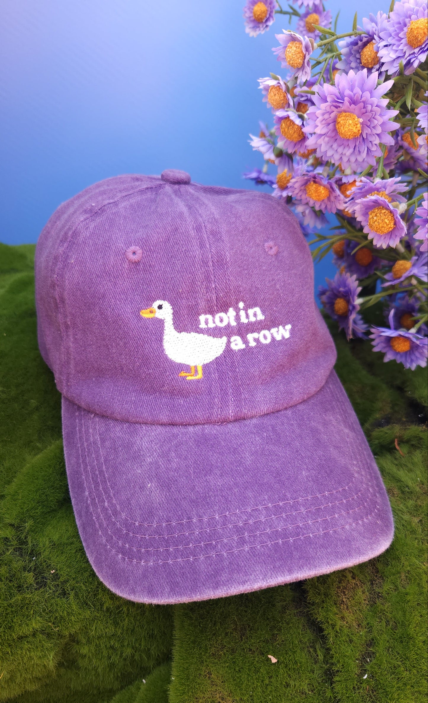 Purple cap with duck design on a green surface with flowers in the background