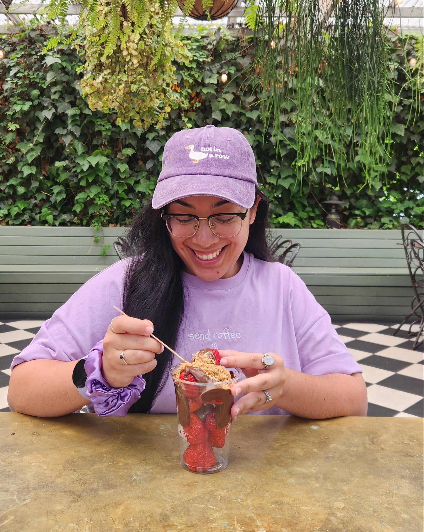 Woman in a purple shirt and cap holding a drink, surrounded by plants and a checkered floor.