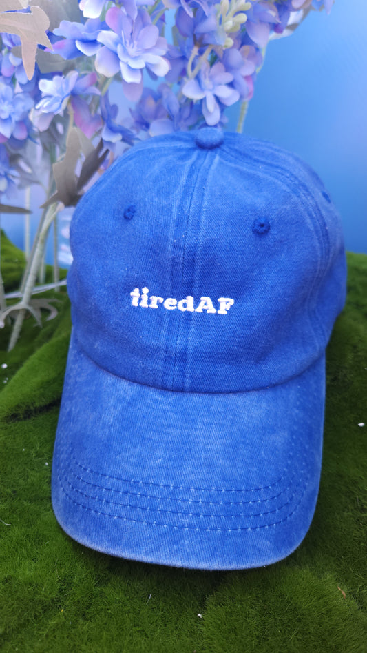 Blue cap with 'tiredAF' text embroidered, on a green surface with purple flowers